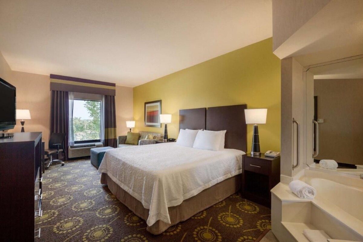 A hotel room with a bed, bedside tables, a desk, a chair, a window, and a bathroom with a bathtub.