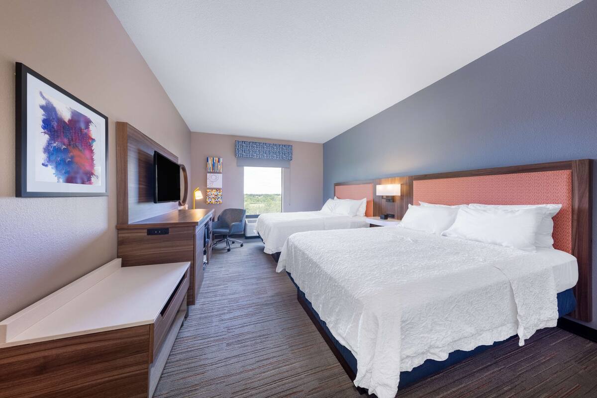 Quarto de hotel com duas camas, uma escrivaninha, televisão e janela com vista.