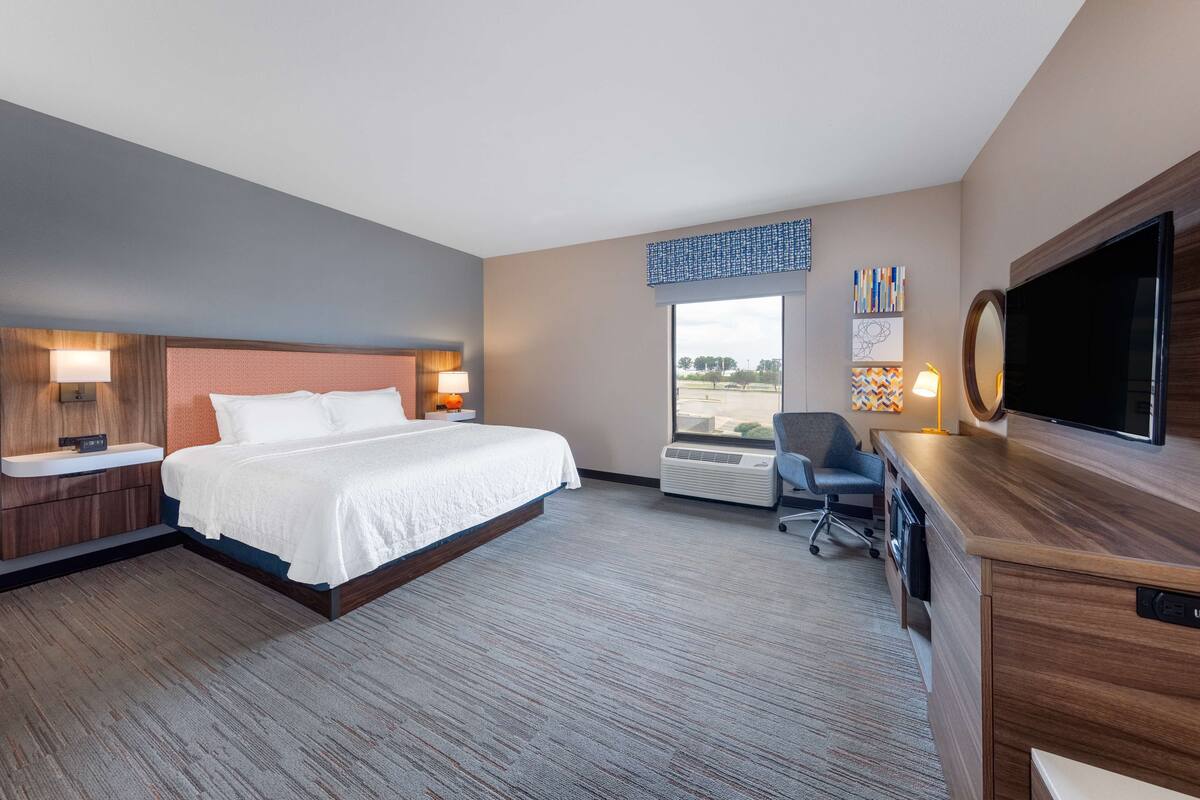 Quarto de hotel com uma cama grande, uma mesa com televisão de tela plana, uma cadeira, um abajur, uma TV montada na parede e uma janela com vista para o exterior.