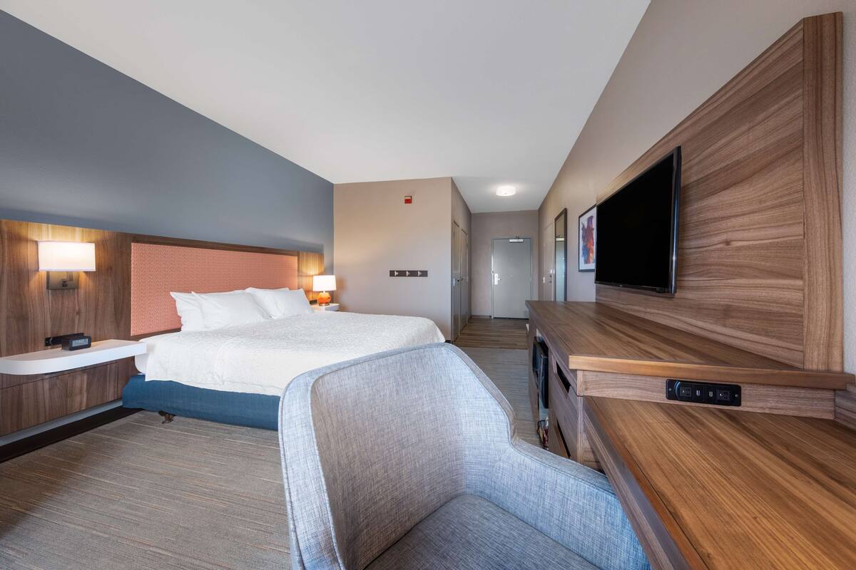 Quarto de hotel com cama, mesas de cabeceira, televisão, poltrona e uma parede de destaque em madeira.