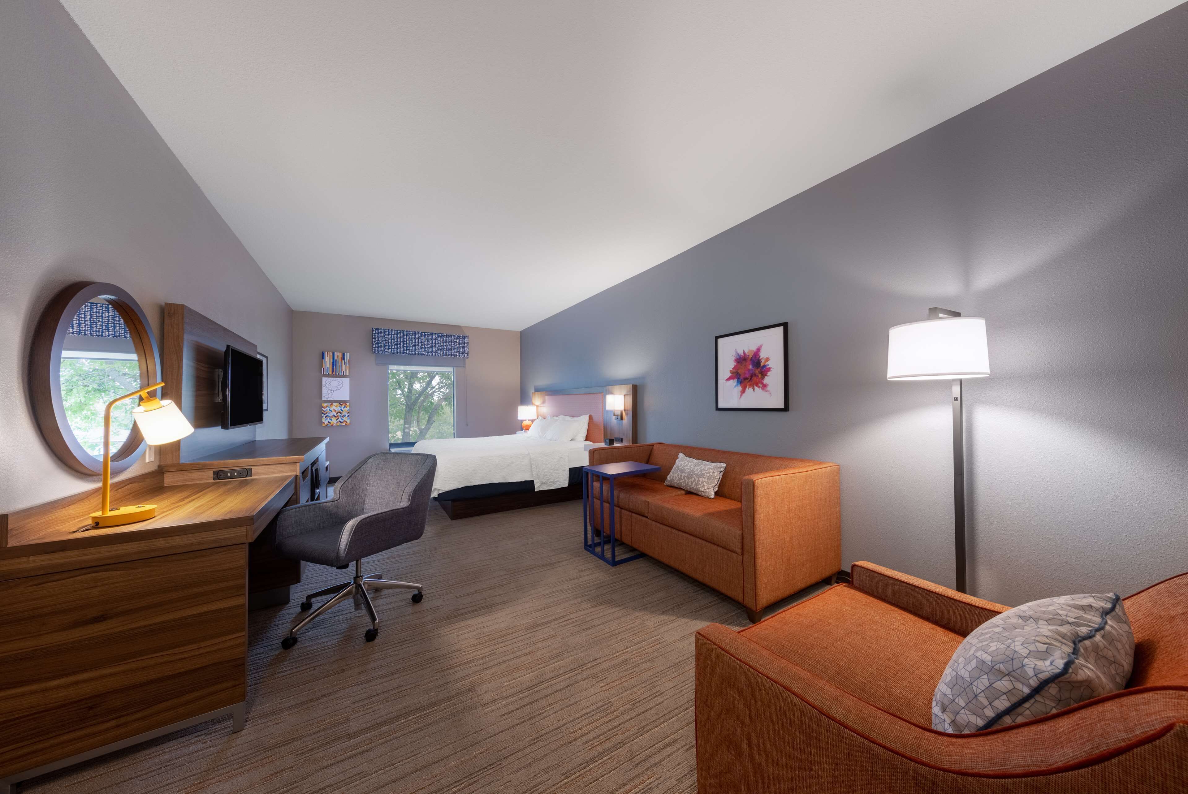 A hotel room with a bed, a desk with a lamp, a chair, an orange sofa, and a standing lamp.