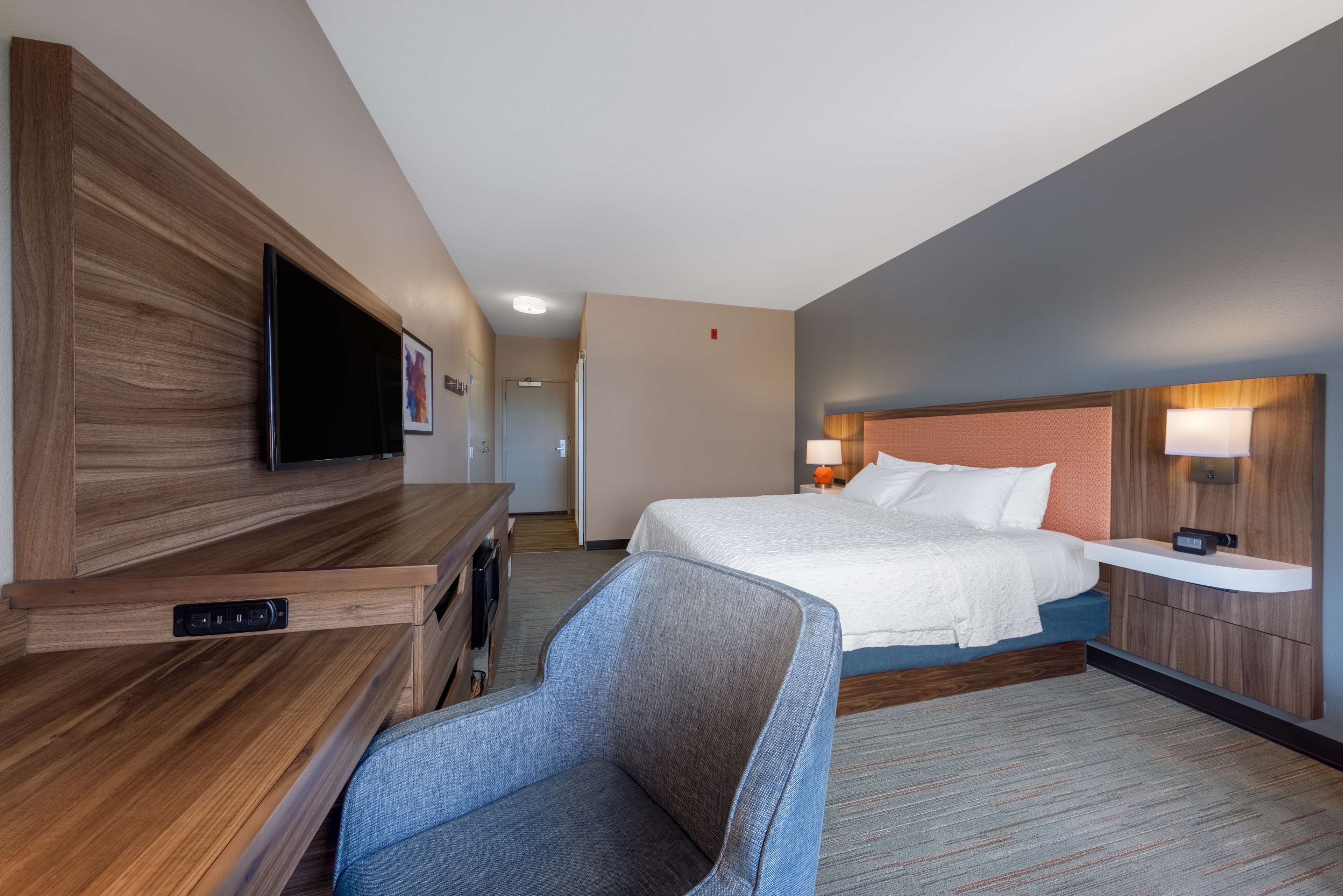 A hotel room with a bed, a desk with a flat-screen TV, a chair, and a bedside table with a lamp.