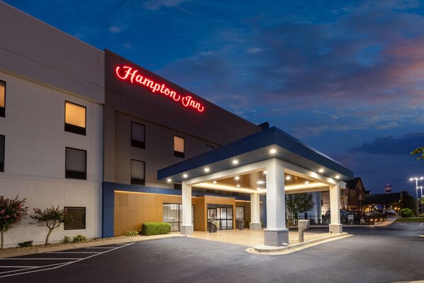 A modern hotel entrance with a prominent red neon sign, a covered outdoor area, and a parking lot.