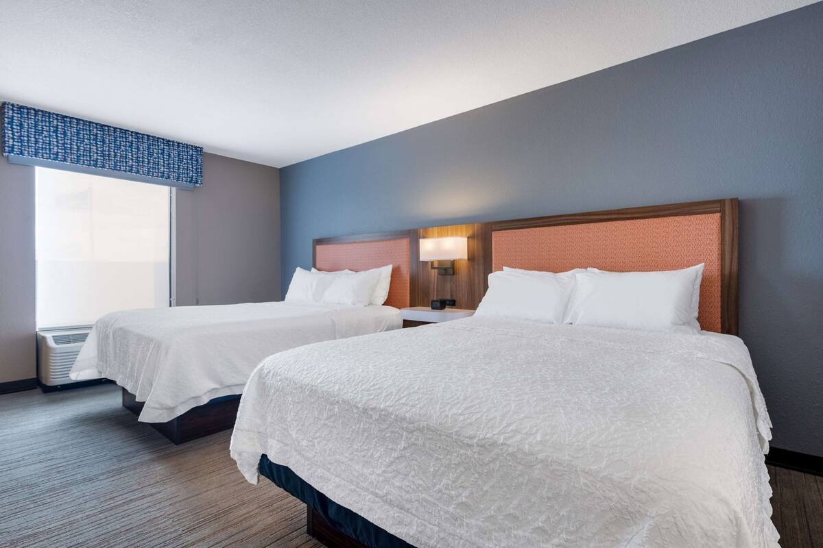 Quarto de hotel com duas camas, janela com persianas, luminária fixada na parede e cabeceira de madeira.
