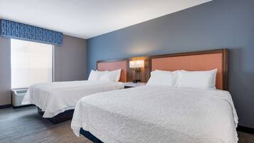 A hotel room with two beds, a window with blinds, a wall-mounted lamp, and a ceiling vent.