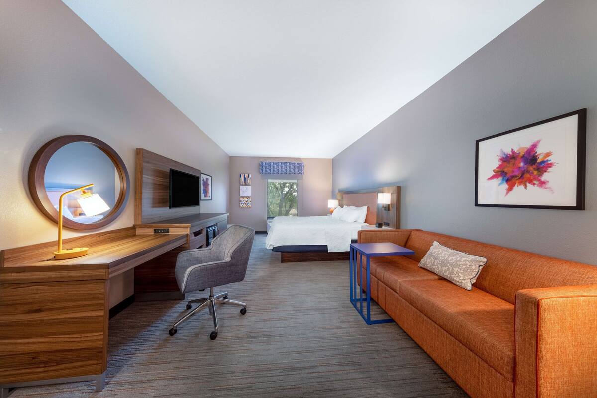 Quarto de hotel com secretária de madeira, espelho redondo, cadeira de escritório cinza, sofá laranja, cama e uma pintura abstrata colorida na parede.