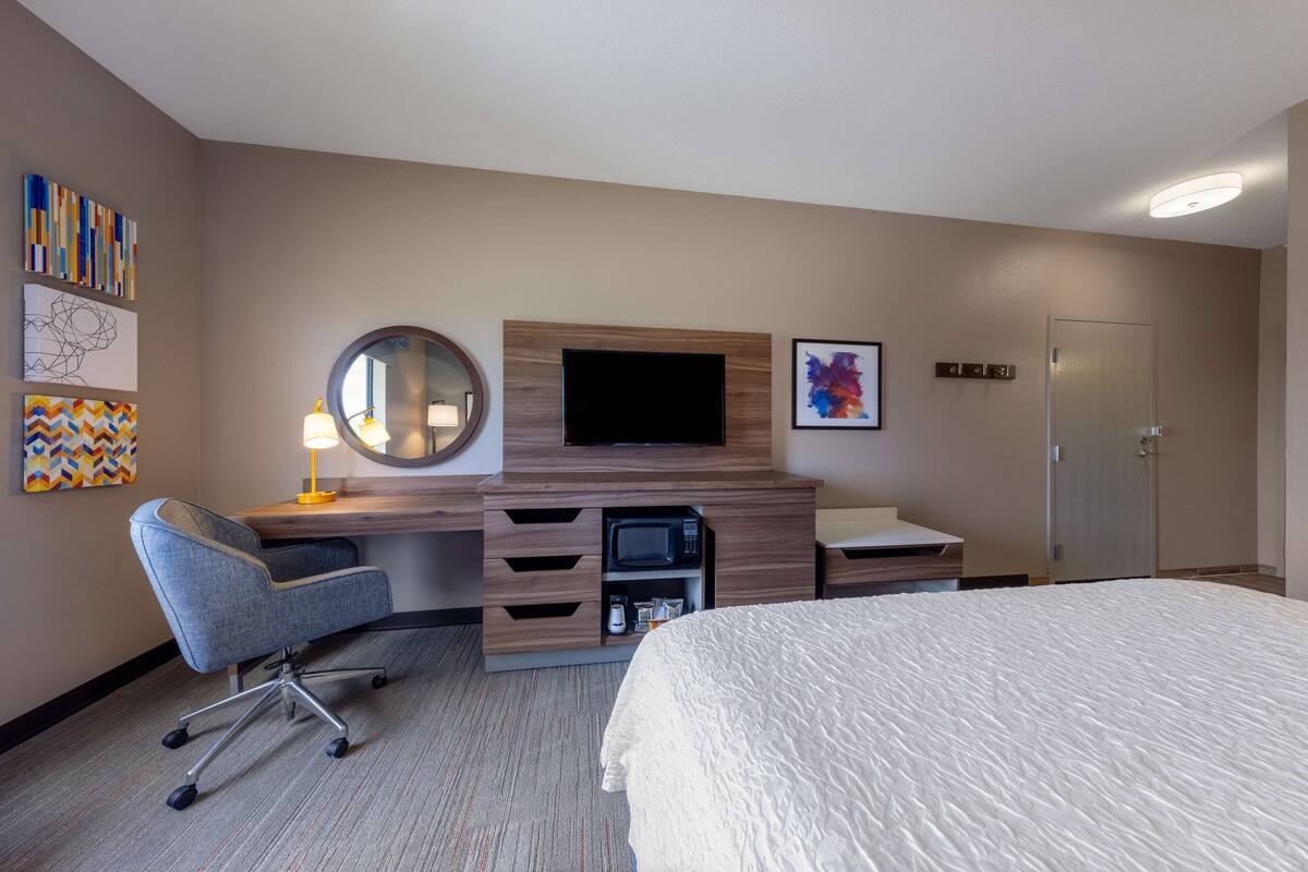 Quarto de hotel com cama, secretária com cadeira, televisão, micro-ondas e porta.