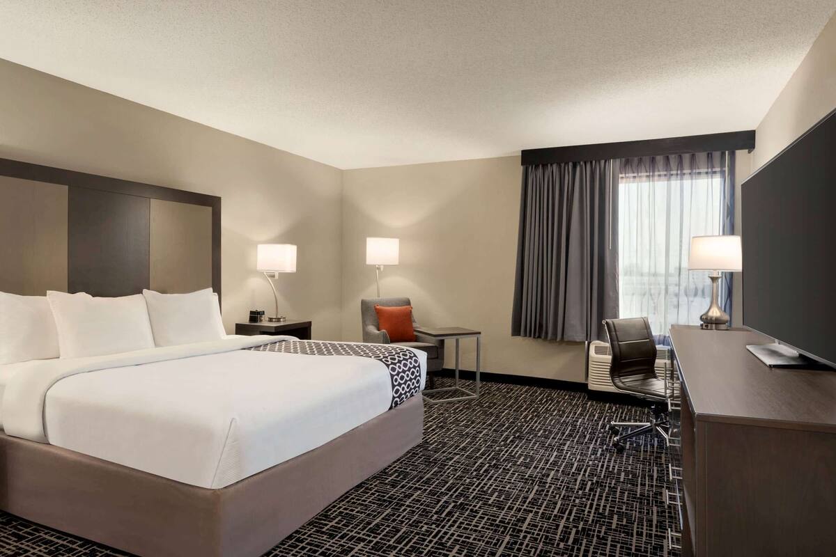 Une chambre d’hôtel avec un grand lit, deux tables de chevet avec des lampes, un bureau avec une chaise et une grande fenêtre avec des rideaux.