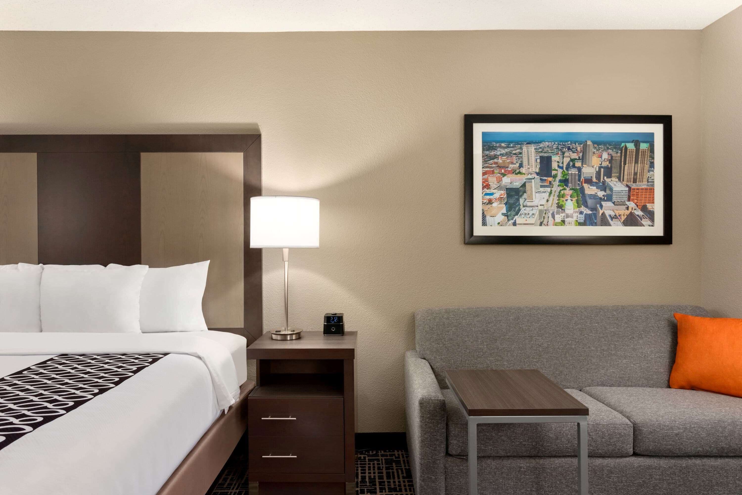 A hotel room with a bed, a nightstand, a lamp, a framed cityscape picture, a gray sofa, and a small table.