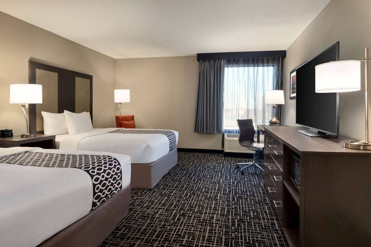 Une chambre d’hôtel avec deux lits, un bureau avec une télévision et une grande fenêtre avec des rideaux.