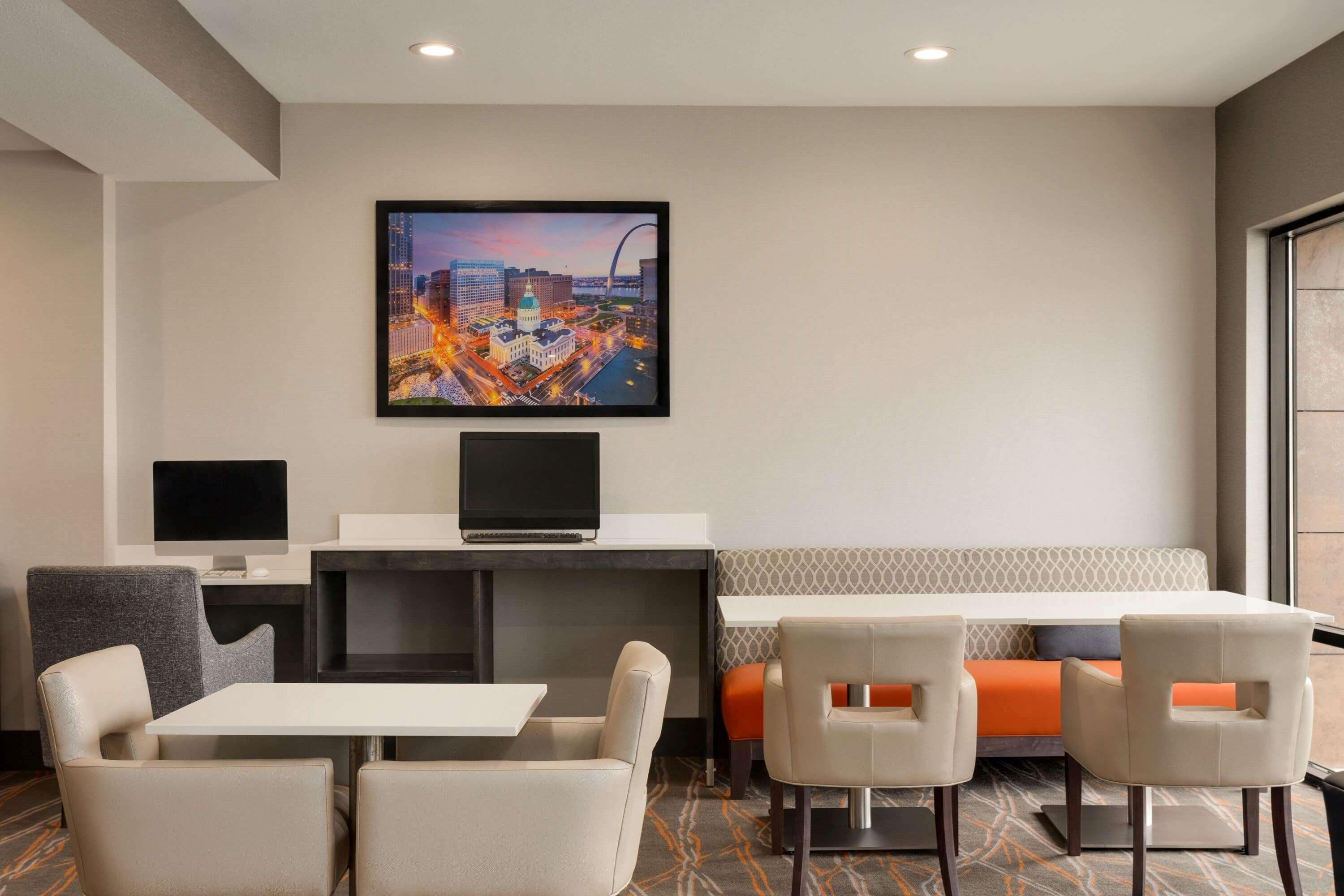 A hotel room with a desk, two chairs, a bench, and a large cityscape painting on the wall.