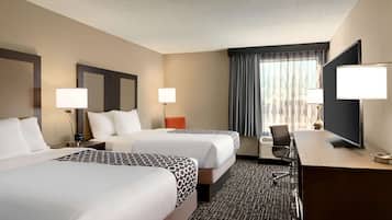 A hotel room with two beds, a desk, a TV, and a large window with curtains.