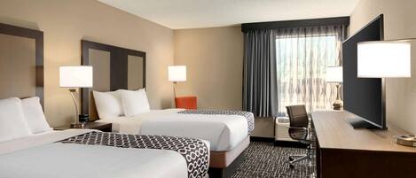 A hotel room with two beds, a desk, a TV, and a large window with curtains.