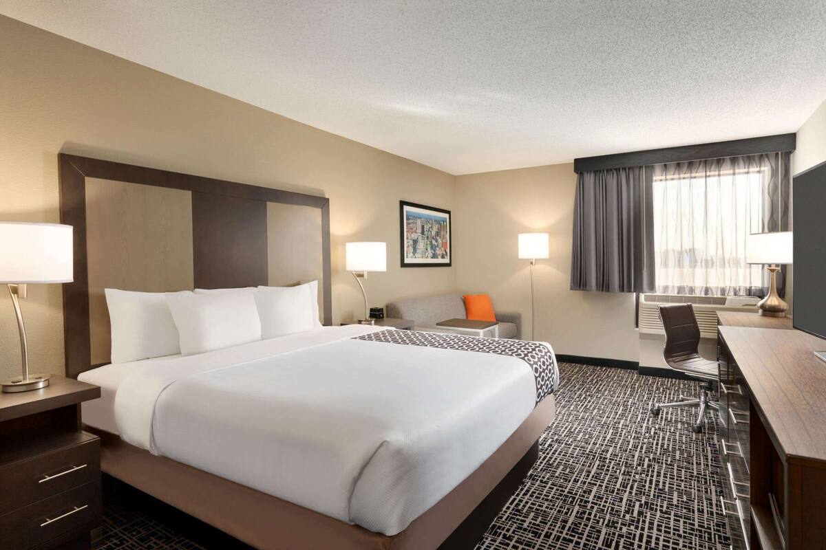 Une chambre d’hôtel avec un grand lit, deux tables de chevet avec des lampes, un bureau avec une chaise, un canapé et une fenêtre avec des rideaux.