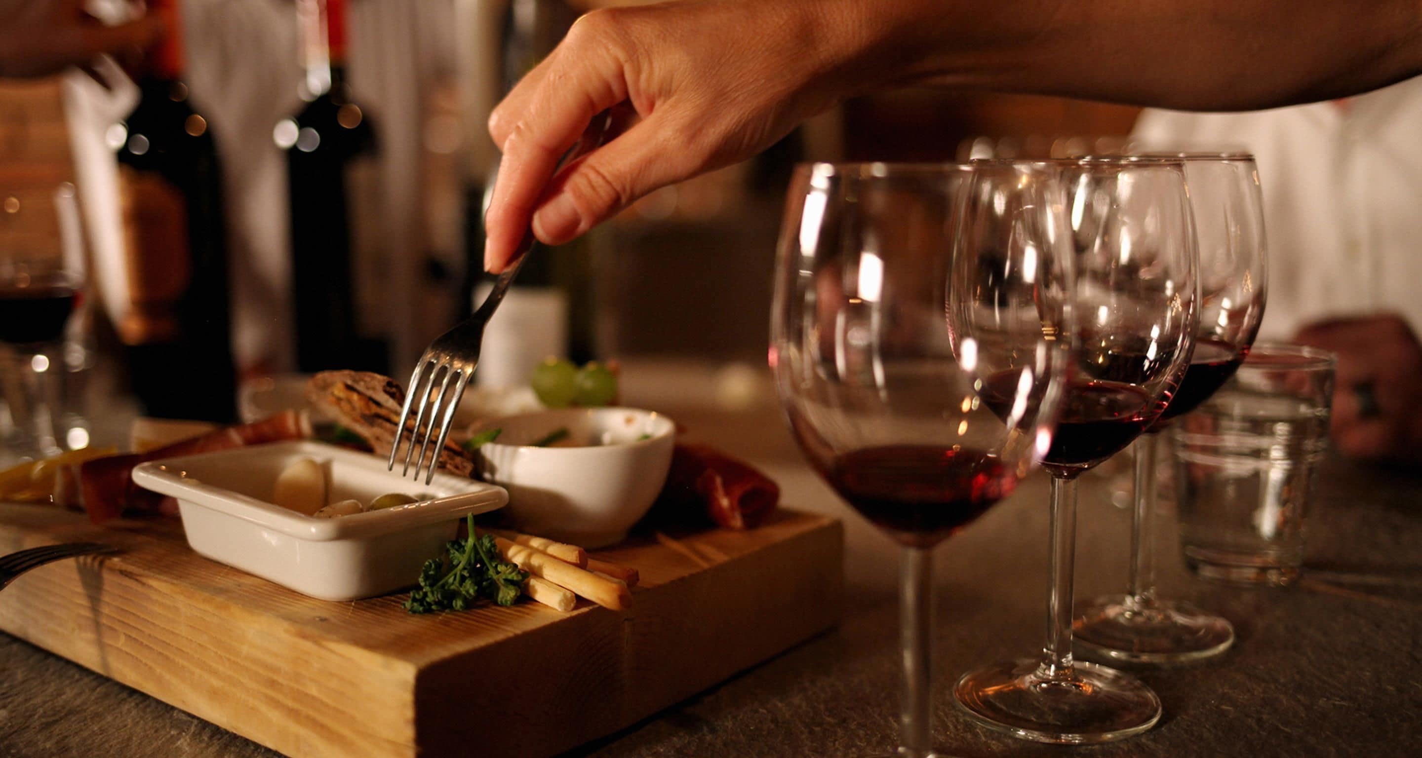 A dining table with a wooden board, a white bowl, and a fork. There are wine glasses and bottles in the background.