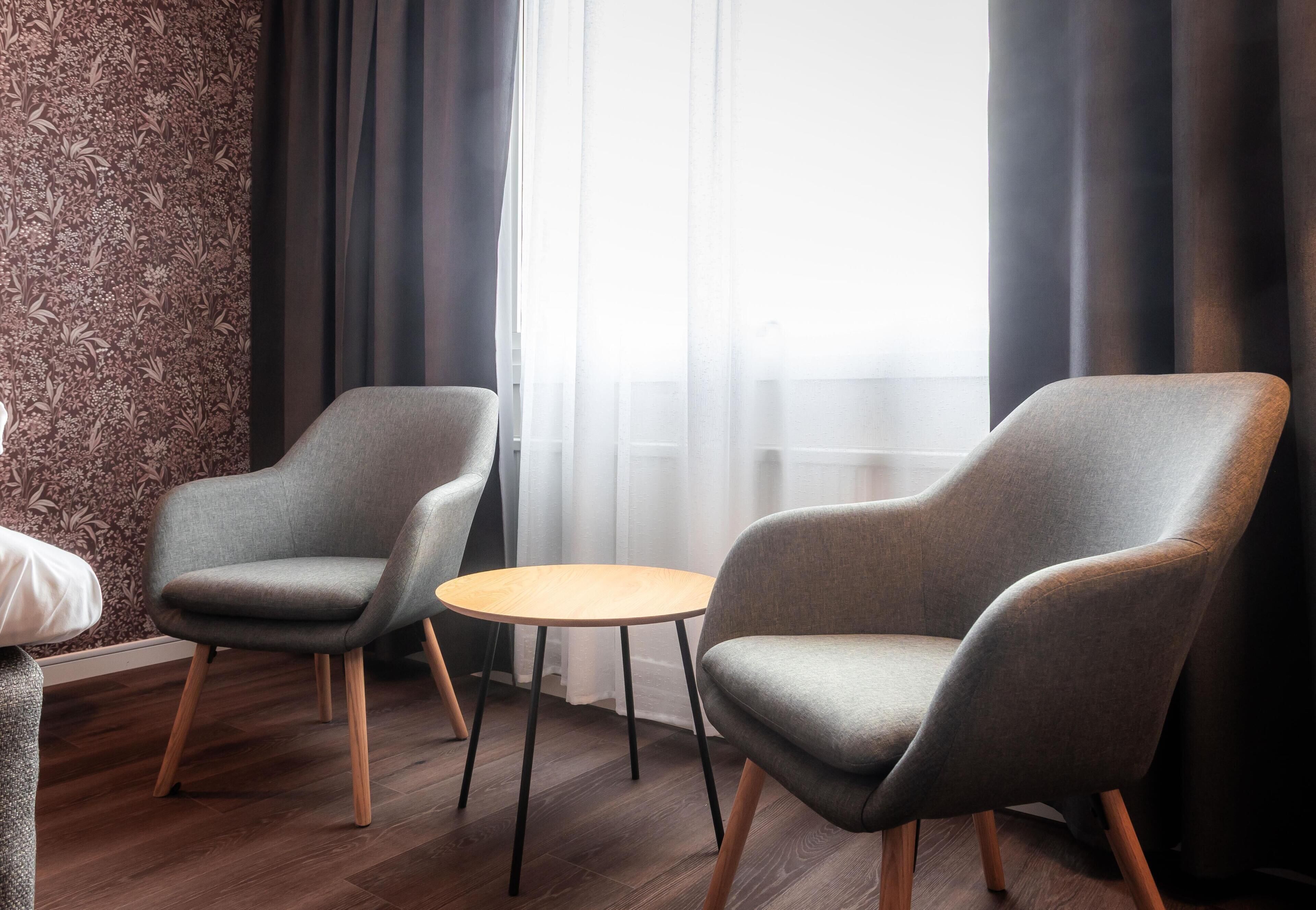 A room with two grey armchairs, a small round table, and a window with sheer curtains.
