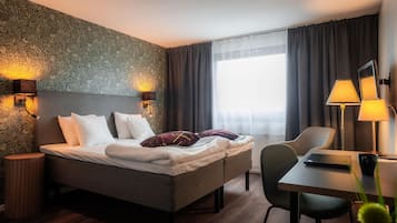 A hotel room with a bed, two bedside lamps, a desk with a chair, and a large window with curtains.
