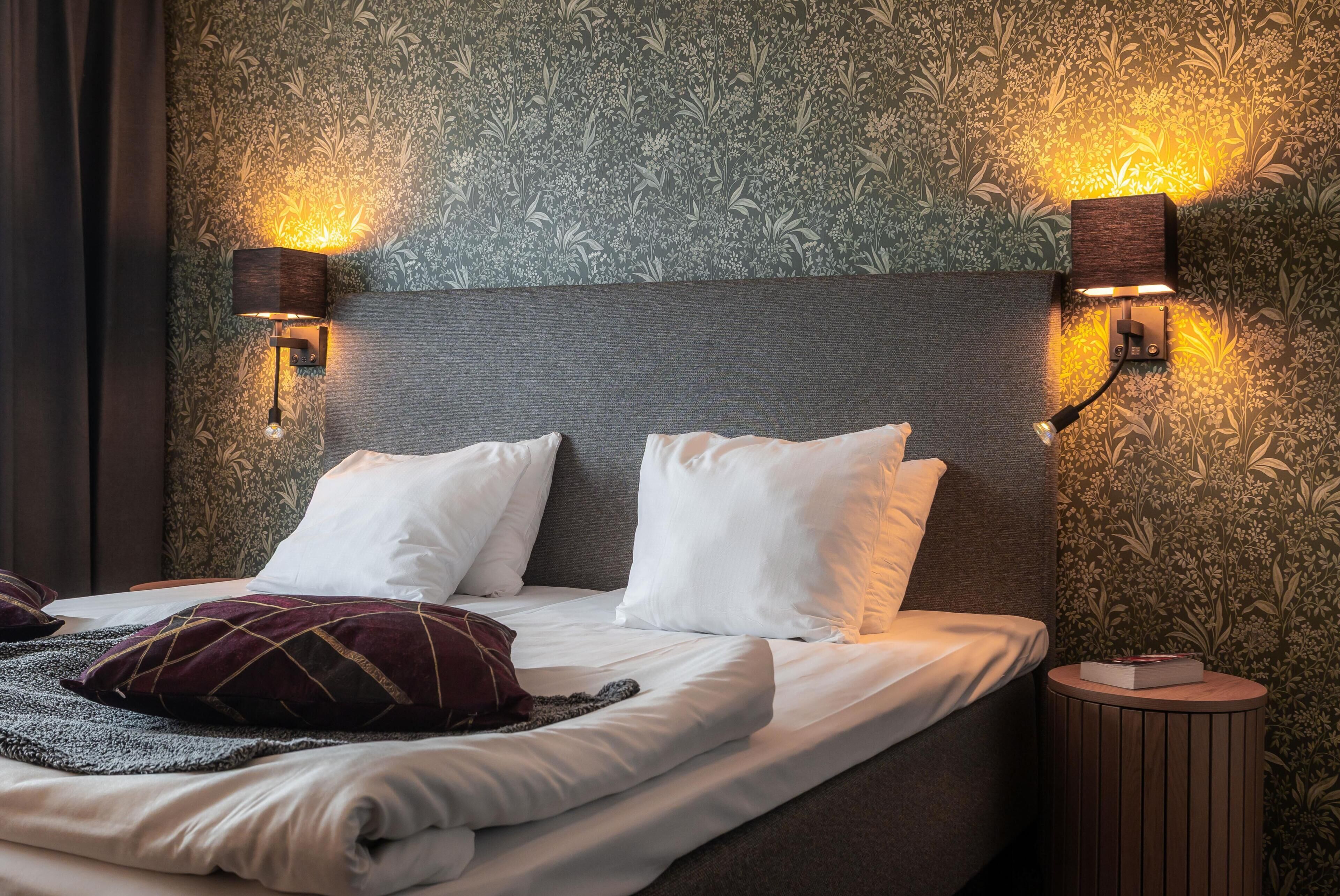Een netjes opgemaakt bed met witte lakens en een donker hoofdbord, twee wandlampen voor het lezen en een rond houten nachtkastje met een boek.