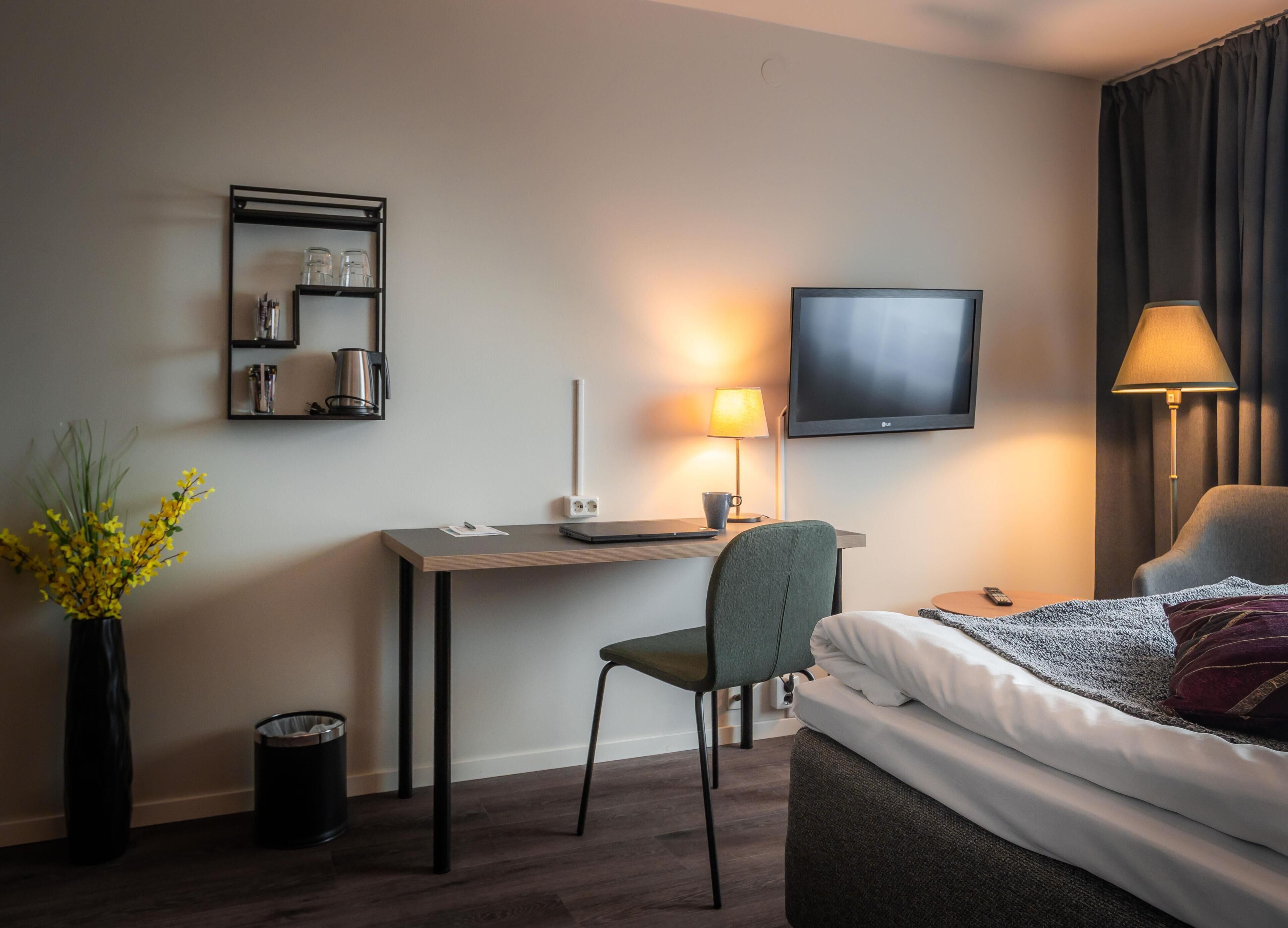 Ett hotellrum med en säng, ett skrivbord, en stol, en TV, en lampa och en liten hylla med diverse föremål.