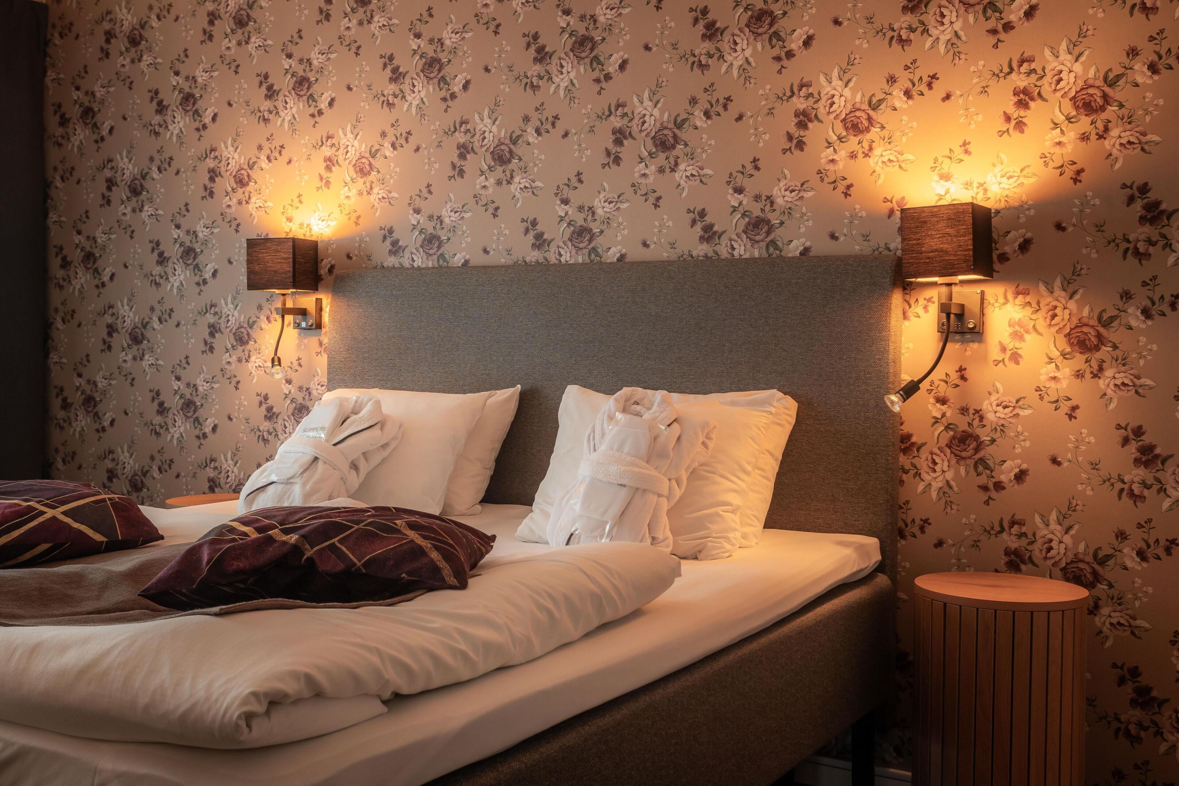 A neatly made bed with white linens and a brown headboard, two wall-mounted lamps, and a round wooden nightstand.