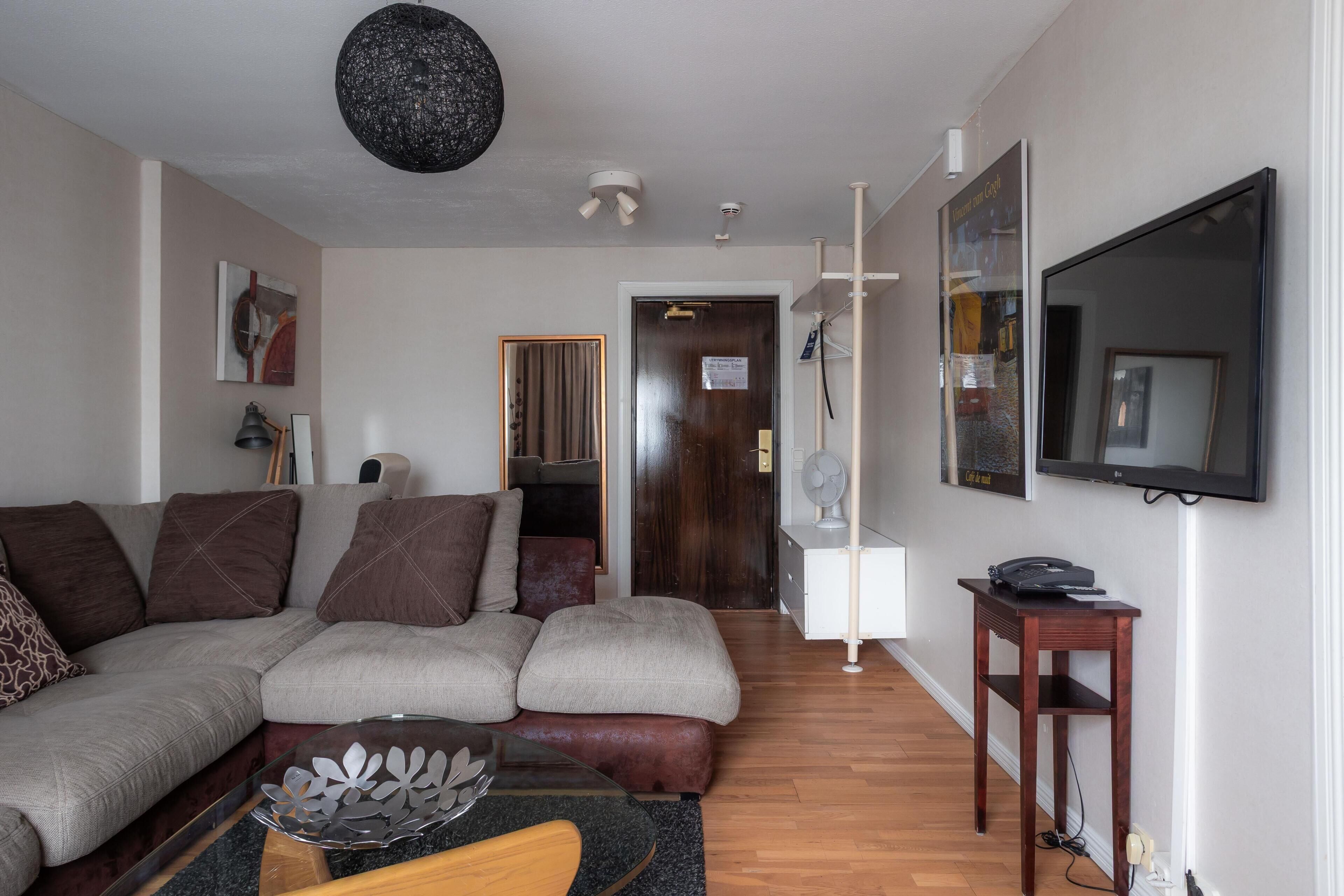 A living room with a sectional sofa, a glass coffee table, a wooden side table, a flat-screen TV, and a door leading to another room.