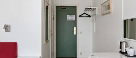Una habitación de hotel con una puerta verde, paredes blancas, una silla roja, una mesita de noche blanca y un aparador blanco con espejo.