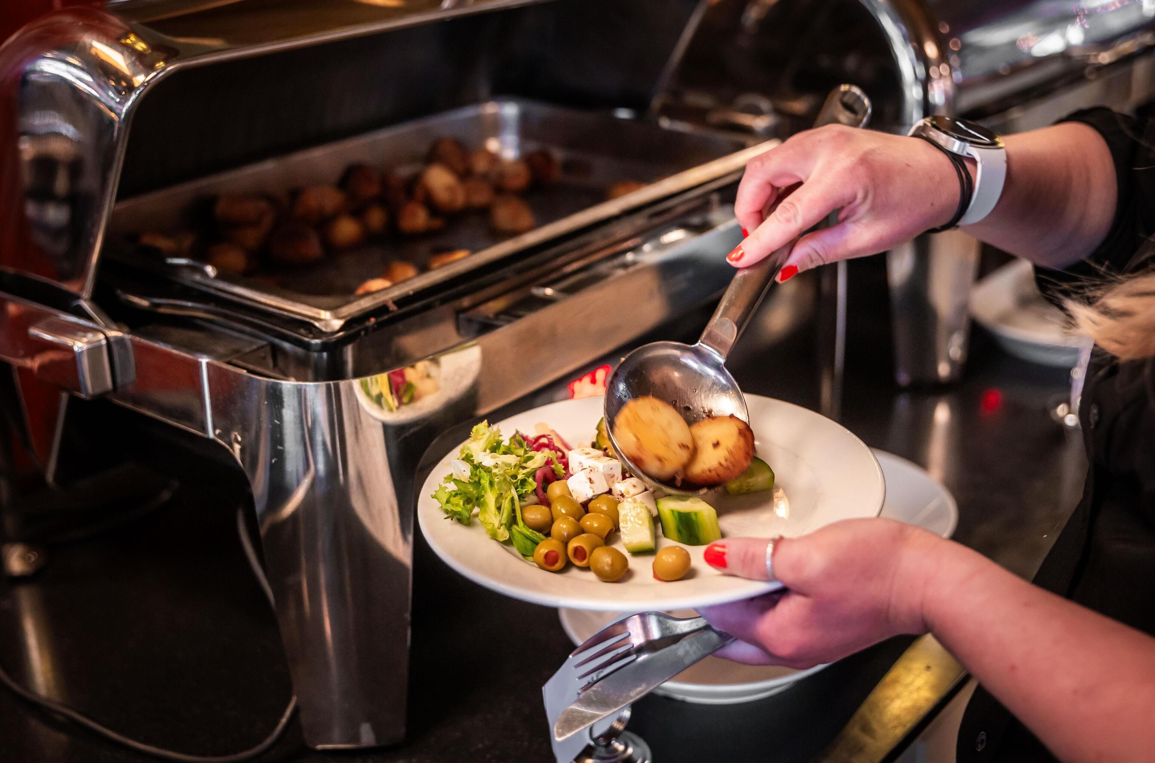 Ein reichhaltiges Buffet mit verschiedenen Speisen, darunter ein Teller mit Oliven, Käse und einem Gebäckstück, sowie eine Person, die vom Grill serviert.