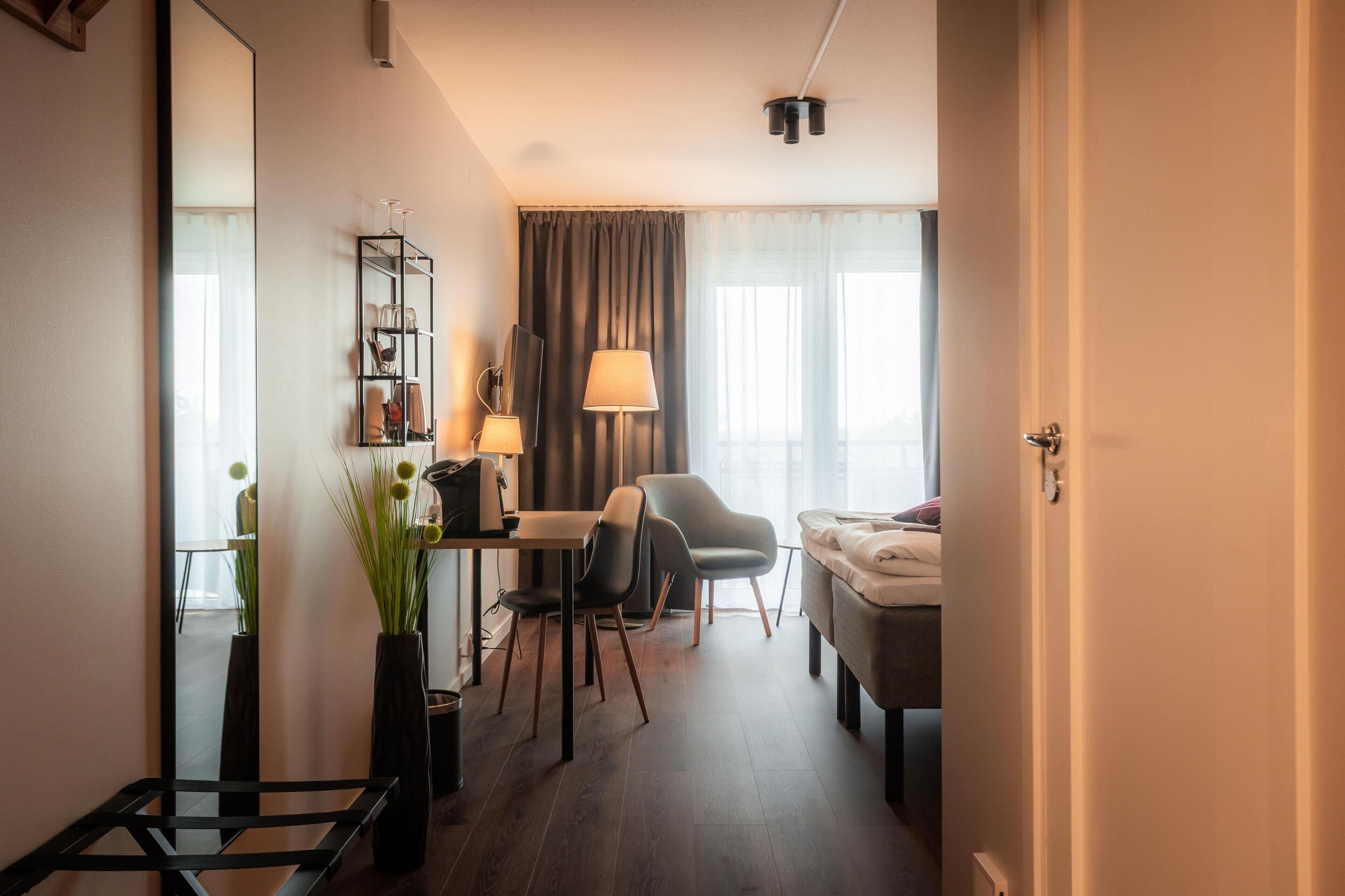 A compact hotel room with a bed, a desk with a chair, a small table, a mirror, a lamp, and a potted plant.