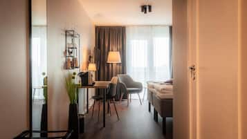 A compact hotel room with a bed, a desk with a chair, a small table, a mirror, a lamp, and a potted plant.
