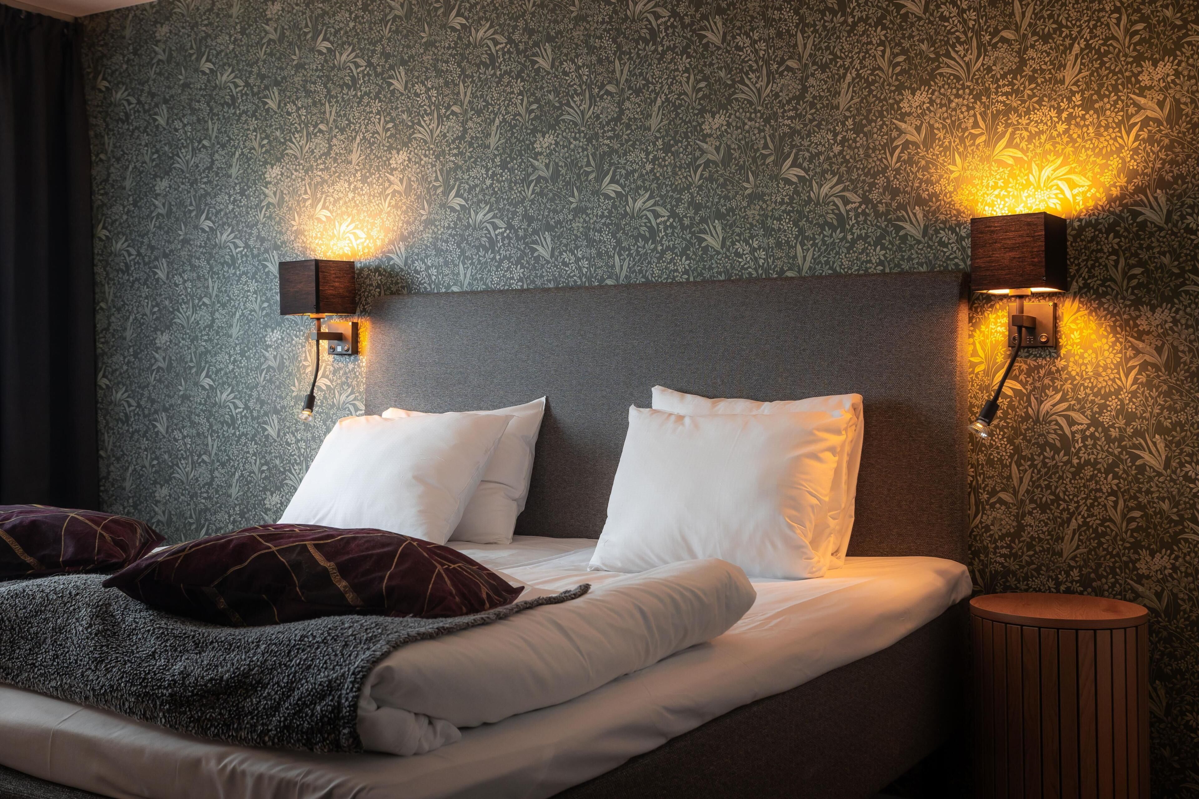 A neatly made bed with white and dark pillows, two wall-mounted light fixtures, and a patterned wallpaper.