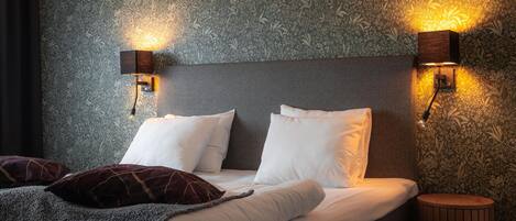 A neatly made bed with white and dark pillows, two wall-mounted light fixtures, and a patterned wallpaper.