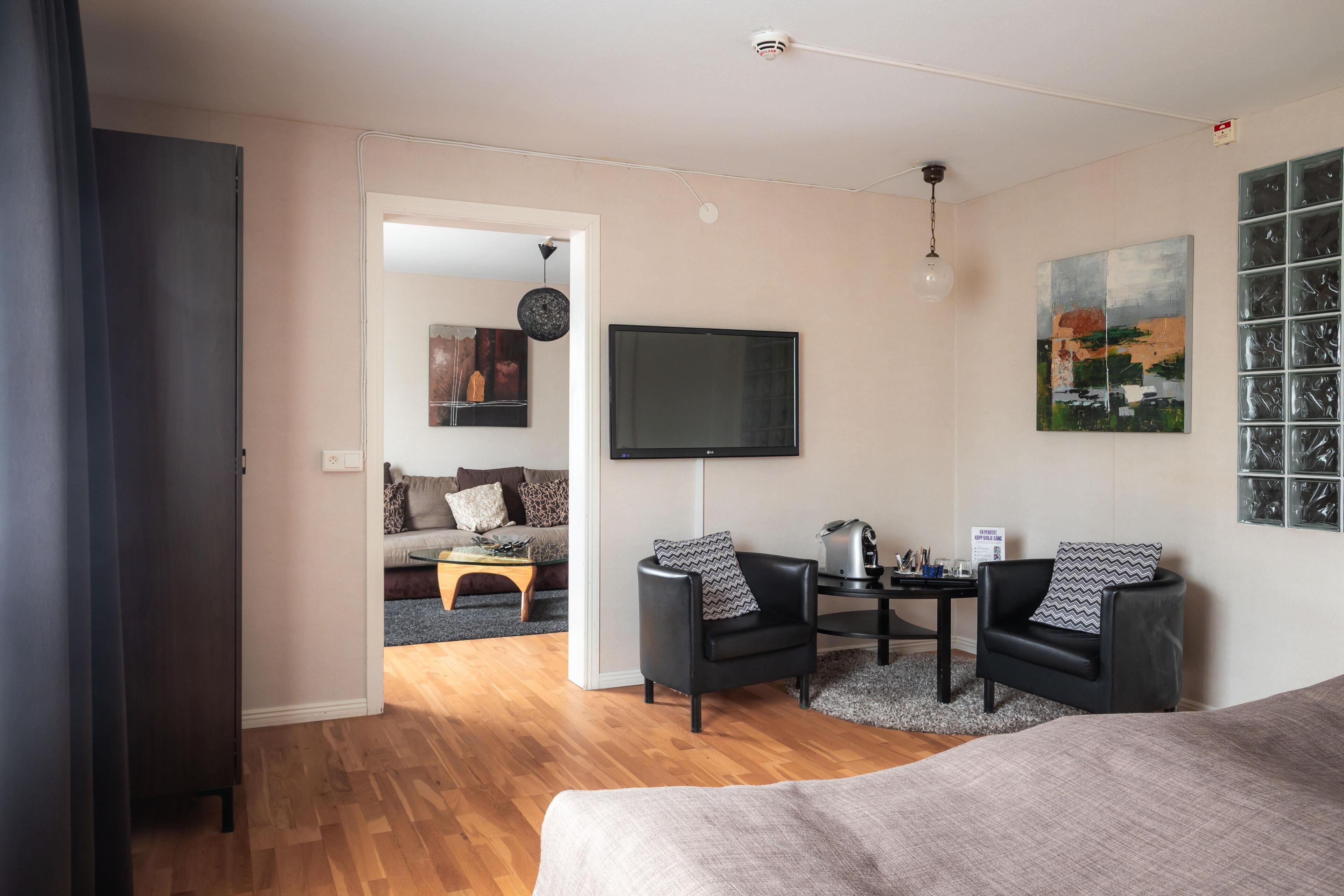 Ein modernes Zimmer mit einem Bett, zwei schwarzen Sesseln, einem Couchtisch, einem Flachbildfernseher und einem Bild an der Wand.