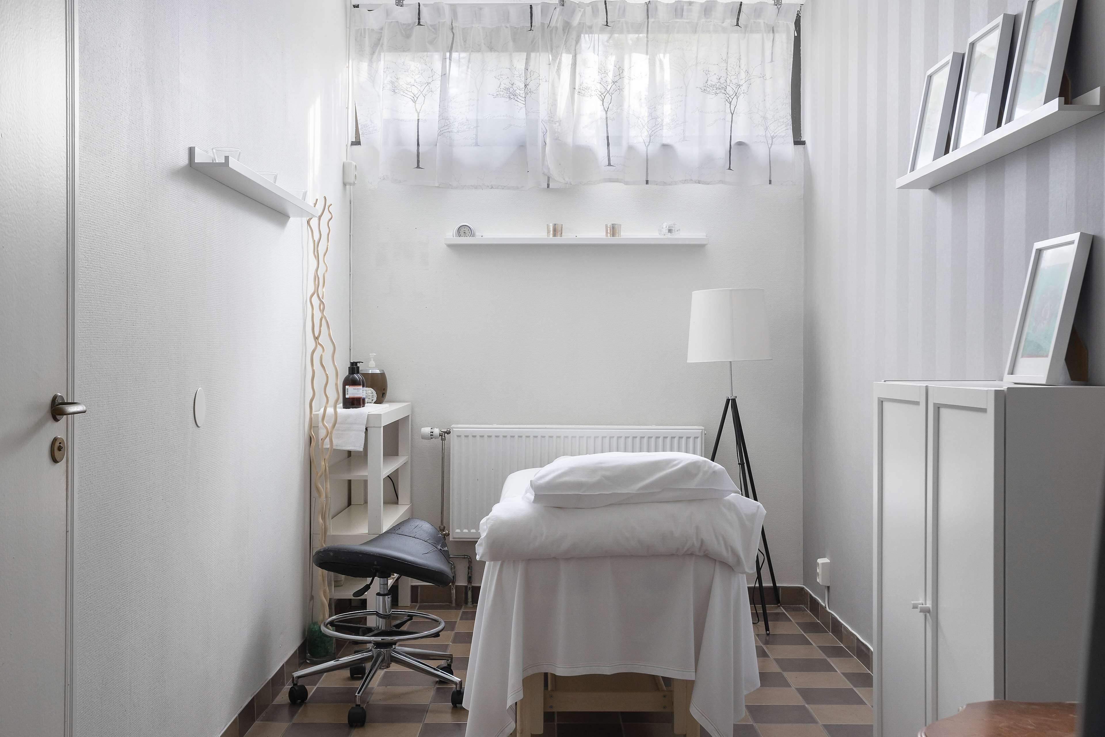 A clinical examination room with a bed, examination table, chair, and shelves with medical supplies.