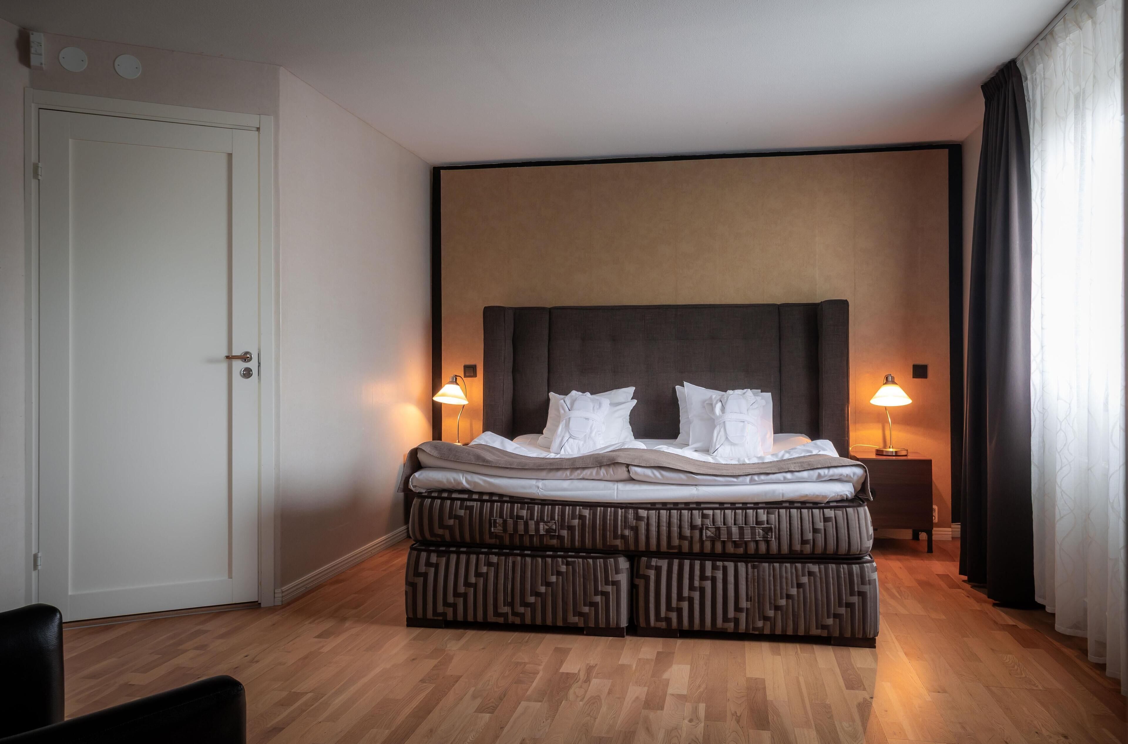 Et hotelværelse med en seng, sengeborde med lamper, en dør og en mønstret skammel.