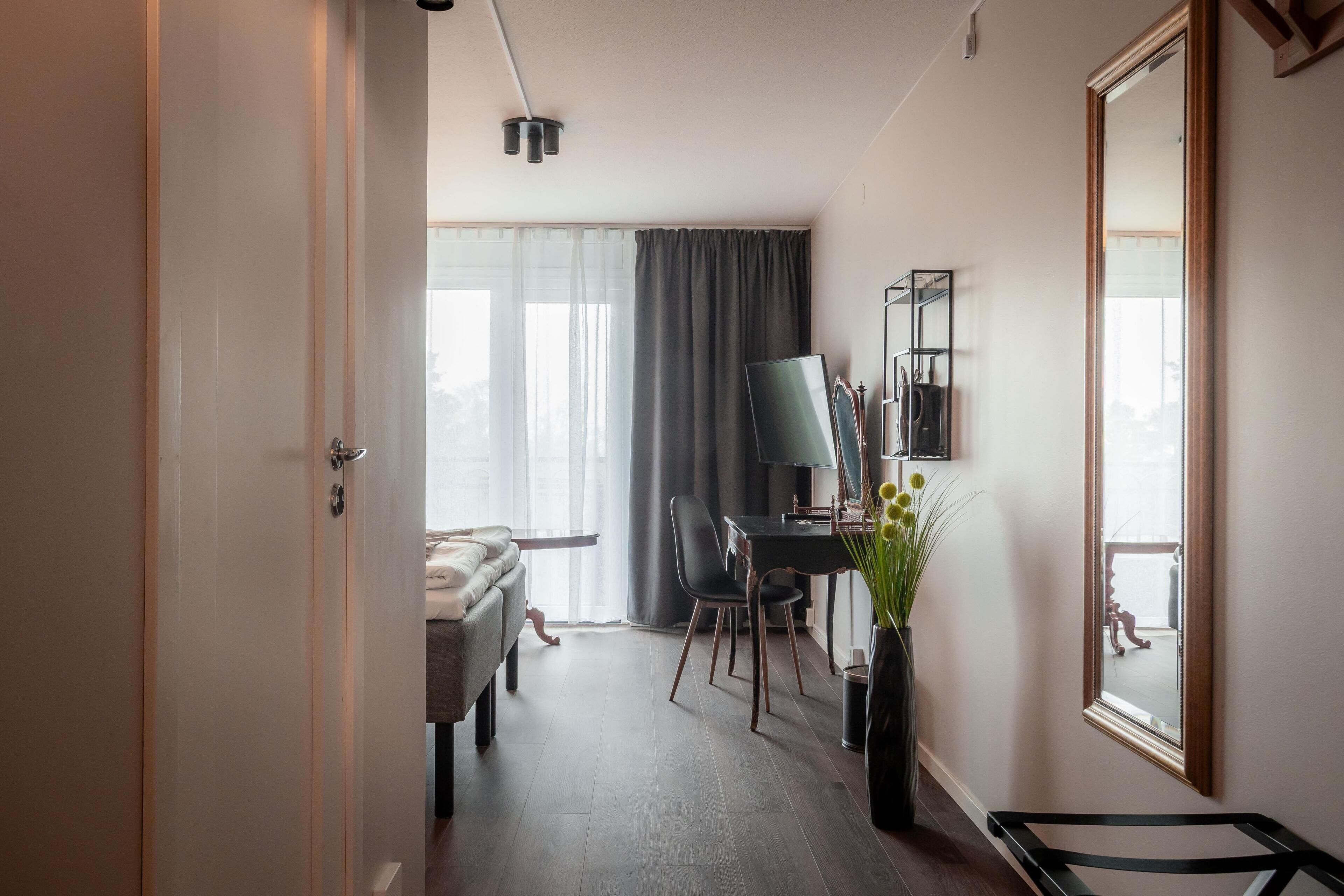 A hotel room with a bed, a desk with a chair, a mirror, and a window with curtains.