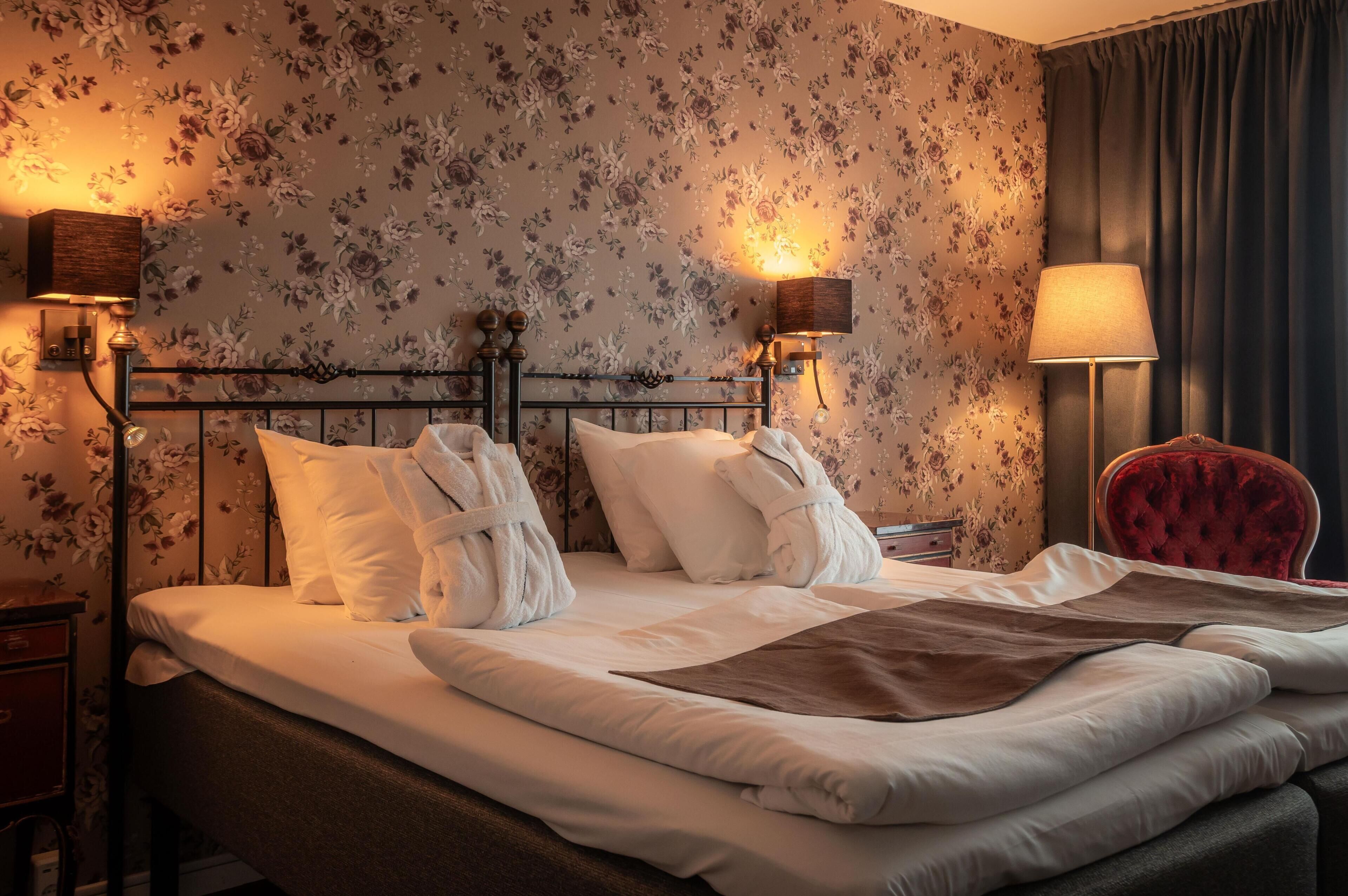 A hotel room with a bed, floral wallpaper, two bedside lamps, a red chair, and a nightstand.