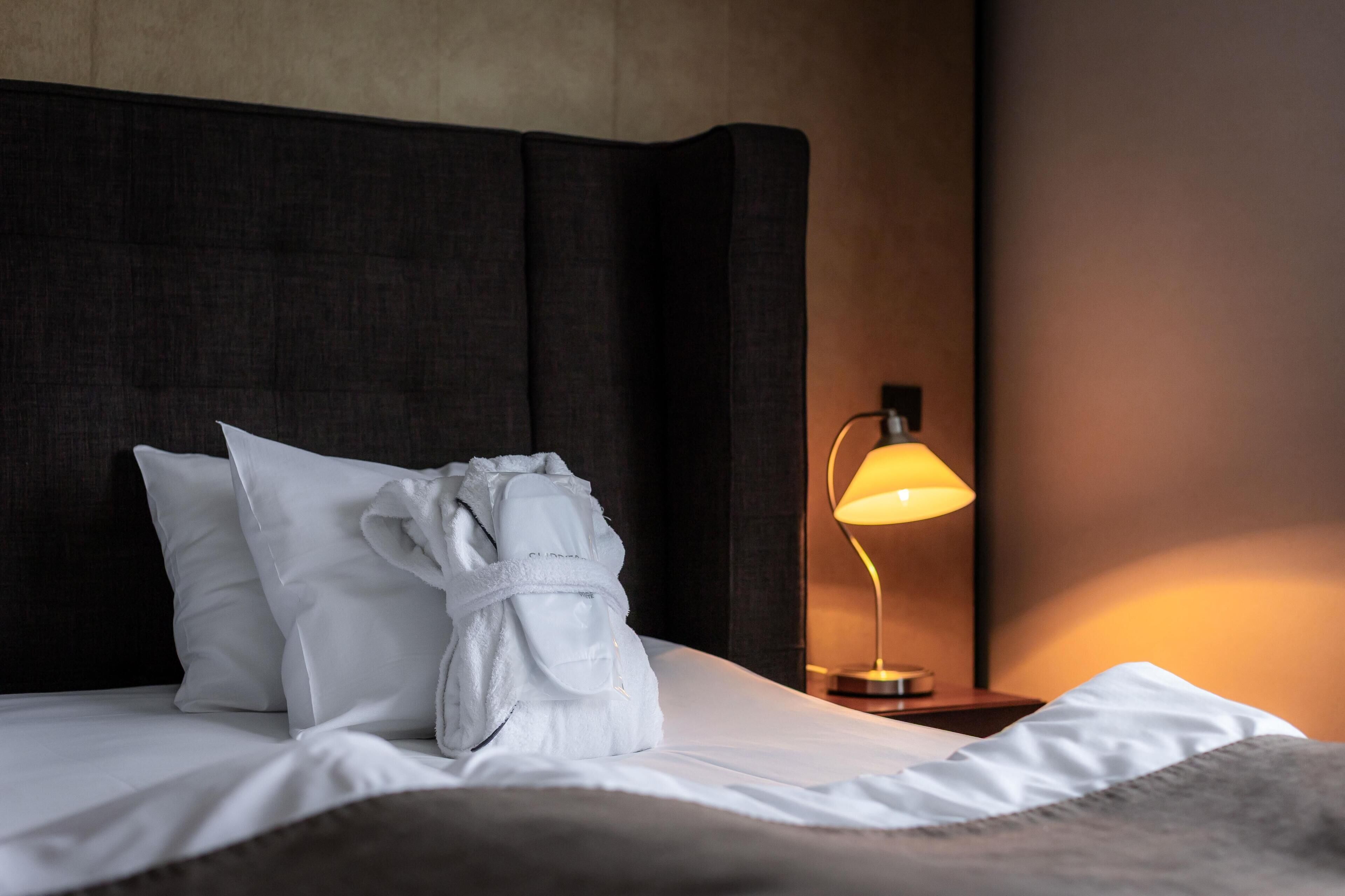 A neatly made bed with white linens, a white bathrobe, and a bedside lamp.