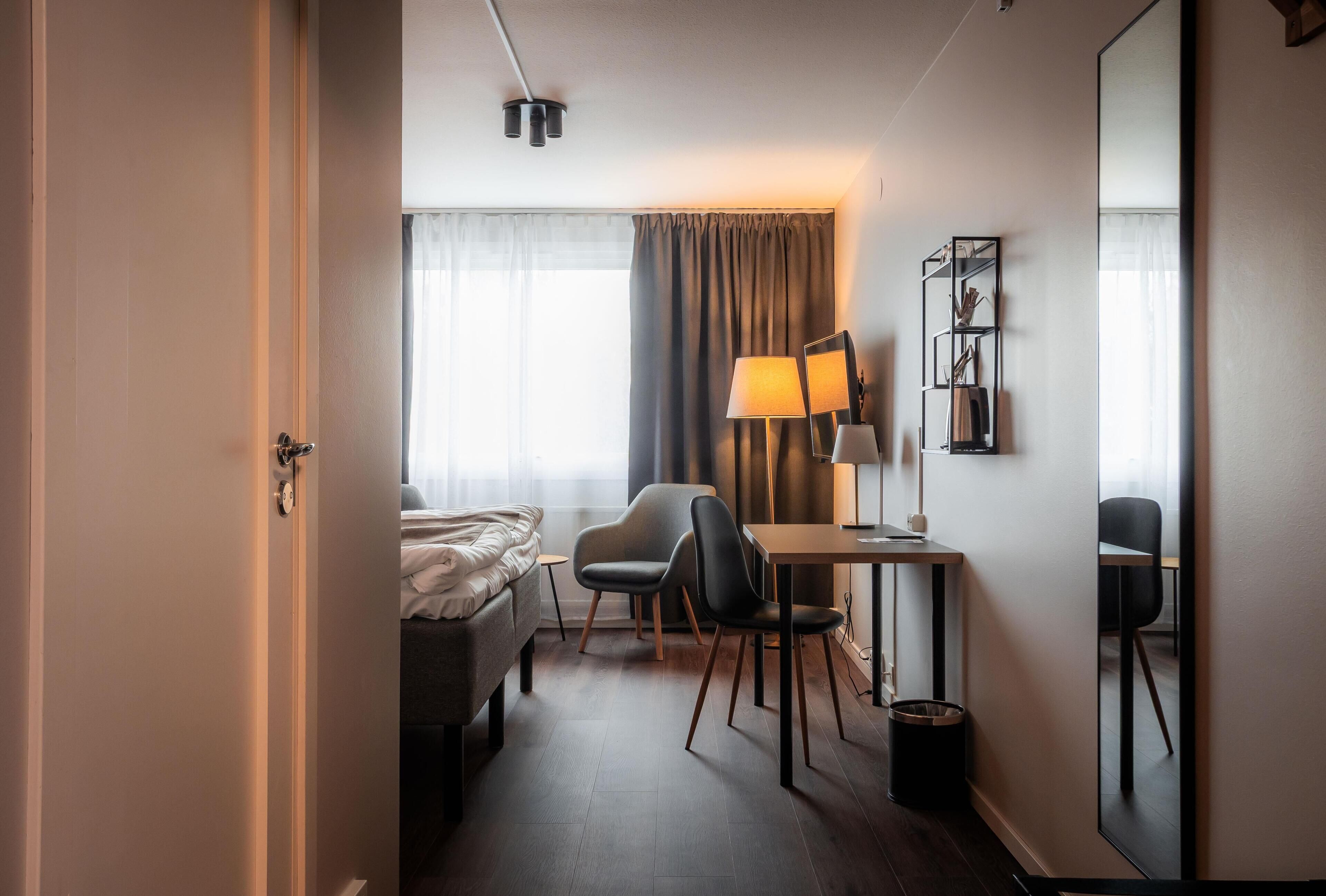 Et hotelværelse med en seng, et skrivebord med lampe, en stol, et lille bord, en skraldespand og et stort spejl.