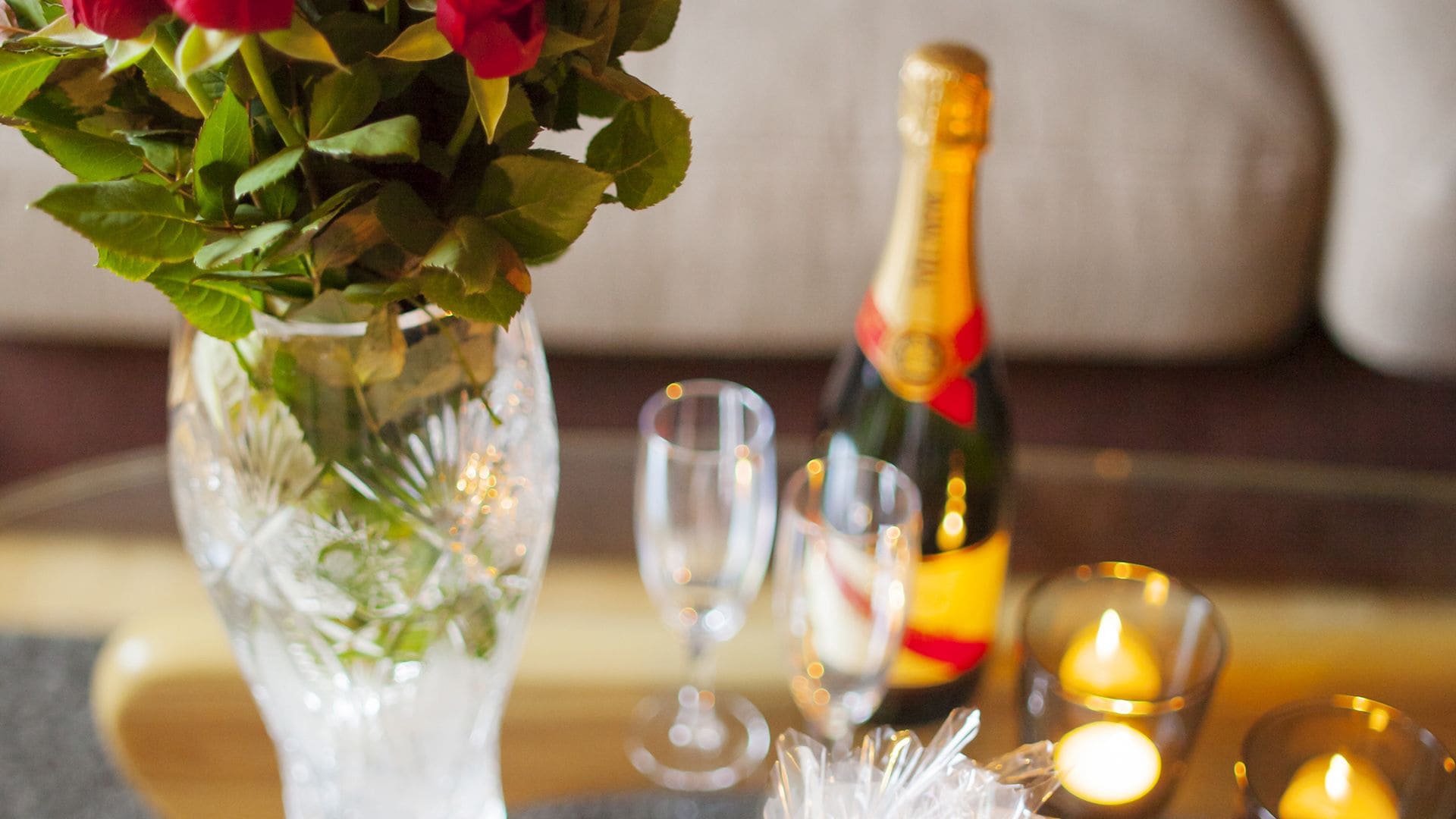 A table with a glass vase containing red roses, a bottle of champagne, and two glasses.