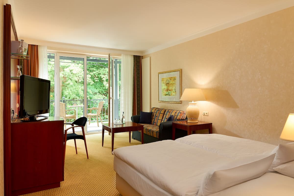 Une chambre d’hôtel comprenant un lit, un bureau avec une télévision, un canapé, une chaise et une table. Il y a une fenêtre donnant sur un paysage verdoyant.