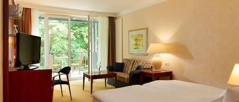 Una habitación de hotel con cama, escritorio con televisor, sofá, sillón y mesa. Hay una ventana con vistas a la vegetación.