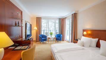 A hotel room with a bed, two blue armchairs, a desk with a lamp, a television, and a window with curtains.