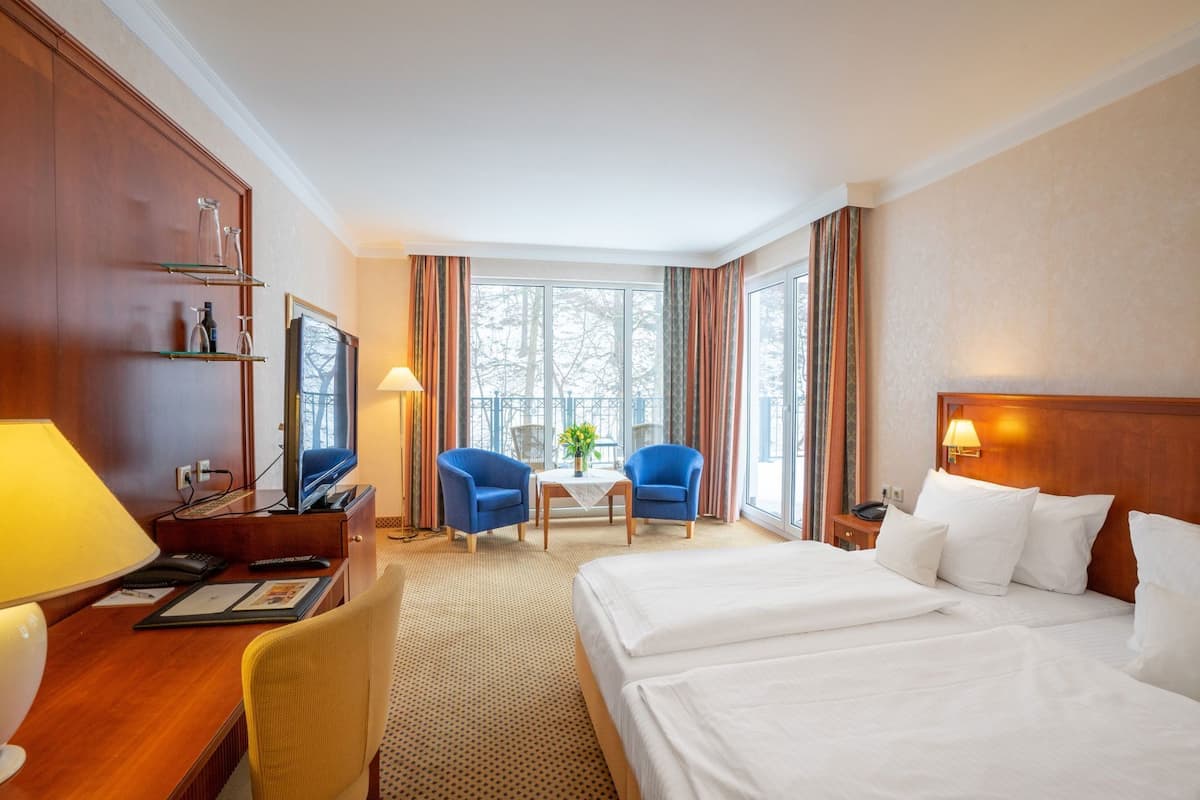 Une chambre d’hôtel comprenant un lit, un bureau avec un ordinateur, une télévision, deux chaises, une petite table et une fenêtre avec des rideaux.