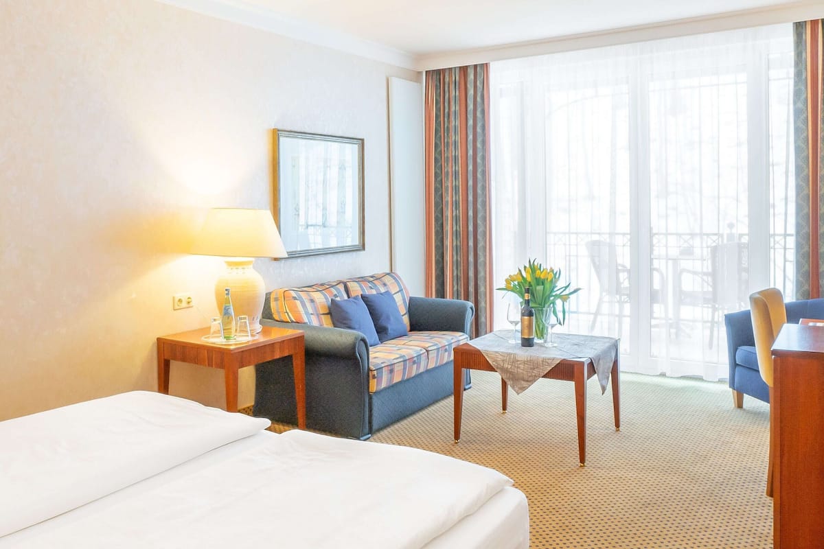 Une chambre d’hôtel avec un lit, un canapé, une table avec un vase, une lampe et une fenêtre avec des rideaux.
