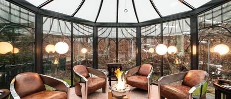 A glass-enclosed seating area with a central fire feature, surrounded by leather chairs and a small table.
