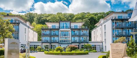A grand hotel entrance with a central glass facade, surrounded by lush greenery and a paved driveway.