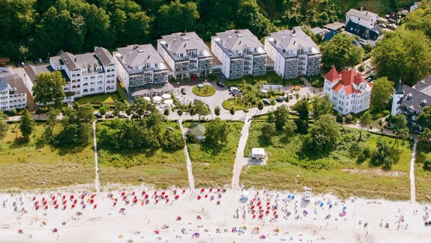 An aerial view of a coastal residential area with multiple apartment buildings, a sandy beach, and a clear turquoise sea. - Grand Hotel Binz (Binz)