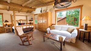 A cozy room with a wooden ceiling, a white sofa, a rocking chair, and a large window with a view of greenery.