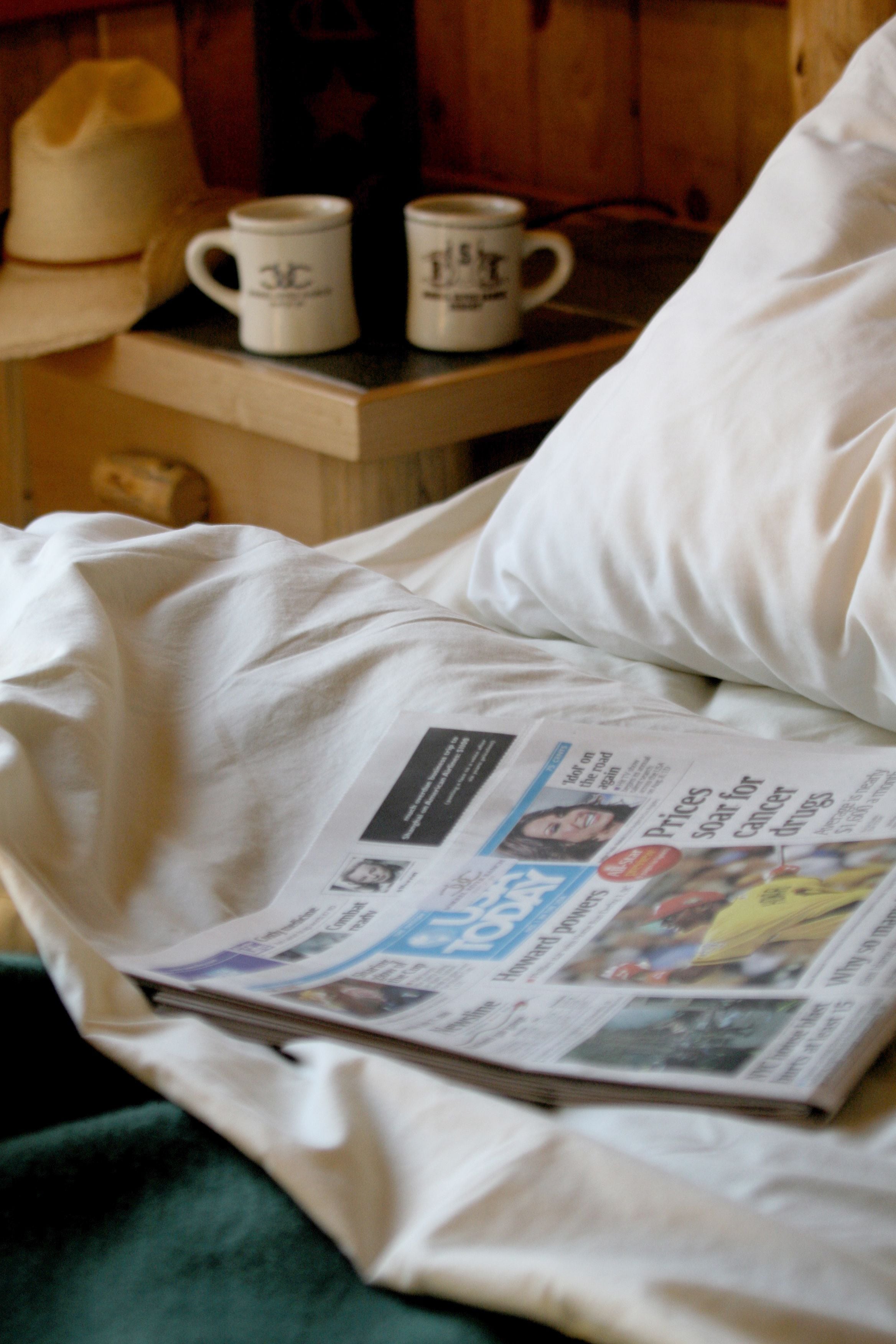 Un lit avec un journal, un oreiller et deux tasses blanches posées sur une table de chevet en bois.