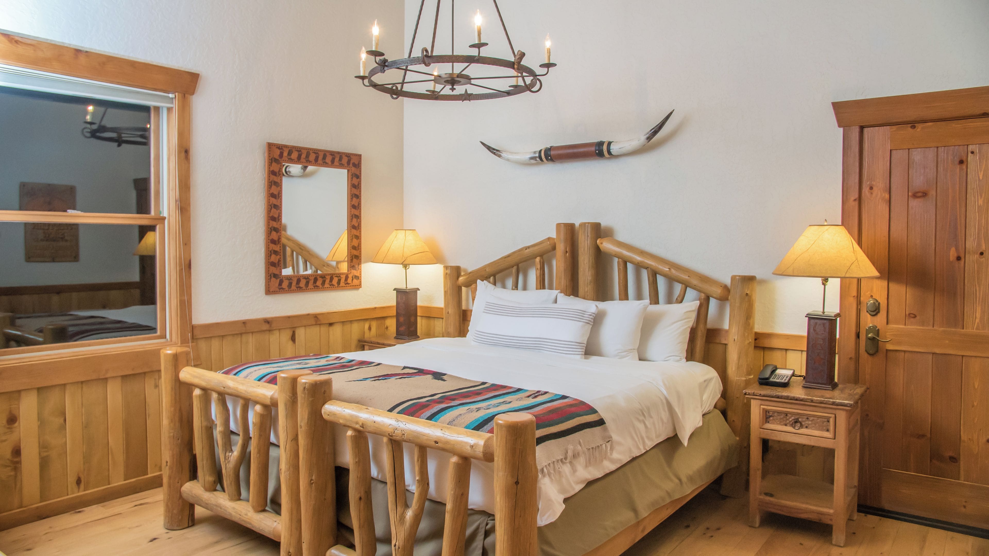 A wooden-framed bed with a colorful blanket, two bedside lamps, a mirror, a chandelier, and a wooden door.